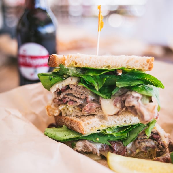 A BBQ brisket sandwich with melted cheese and green lettuce, served with a pickle spear and beer.