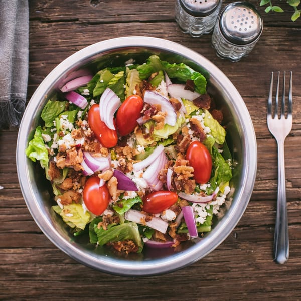 Top-down view of a salad with lettuce, tomatoes, red onion, feta, and bacon bits in a metal bowl.