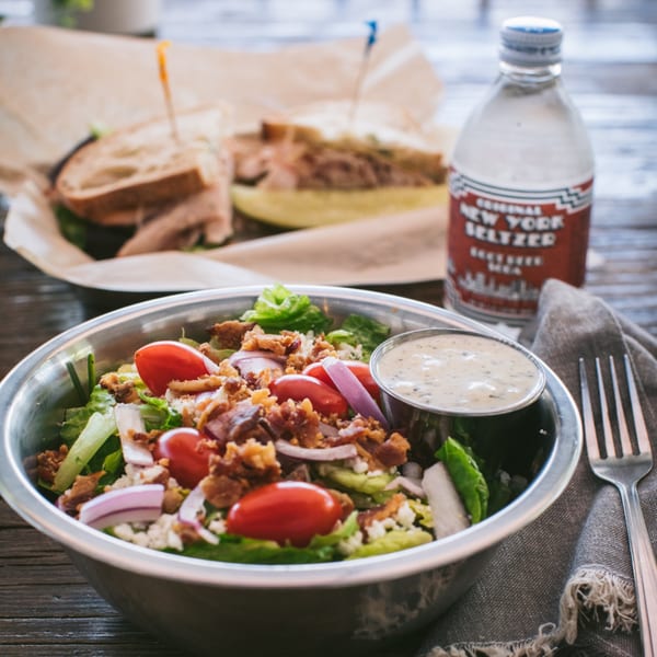 A BBQ salad with tomatoes, bacon, and onions, with a sandwich and seltzer in the background.