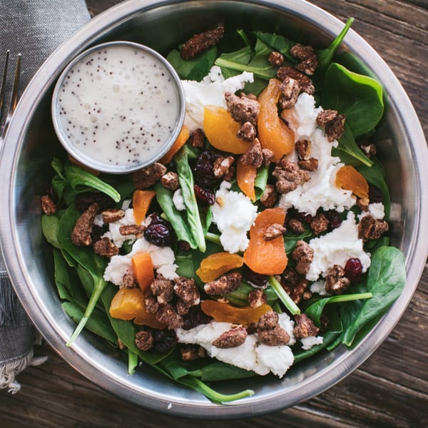 Spinach salad with goat cheese, dried apricots, candied pecans, and poppy seed dressing in a metal bowl.