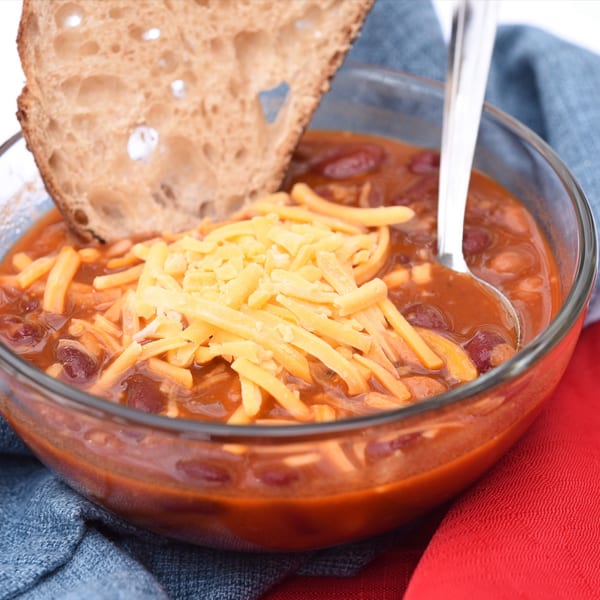 A bowl of American Way Smokehouse chili topped with shredded cheese and a slice of bread.