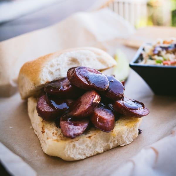American Way Smokehouse BBQ sausage sandwich on a bun with a side dish.