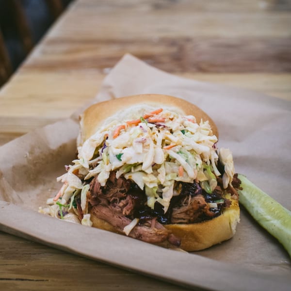 American Way Smokehouse pulled pork sandwich with coleslaw and a pickle spear on a wooden table.