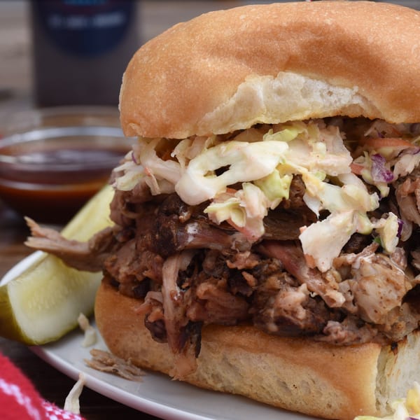 Close-up of a pulled pork sandwich with coleslaw and a pickle spear on a white plate.