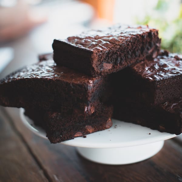 Three rich, fudgy brownies with chocolate chips and a glossy top are stacked on a white cake stand.