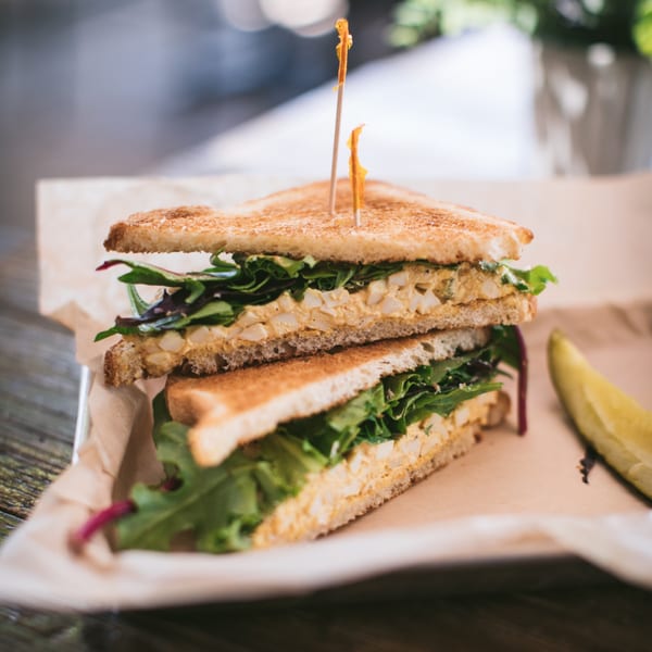 A toasted sandwich with egg salad and greens, held by two picks, served with a pickle spear.